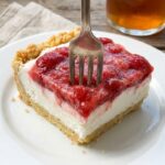 A close-up side view of a square slice of strawberry frozen dessert showing the crust, cream, and berry layers.