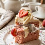 A close-up of a fork cutting into a moist slice of pink strawberry cake on a floral plate.