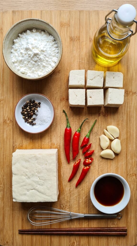 A flat lay showing a block of tofu, cornstarch, chilies, garlic, and spices on a bamboo board