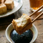 A close-up of chopsticks holding a piece of fried tofu with a bite taken out, showing the soft inside.