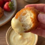 A close-up of a hand dipping a fried fritter into lemon mousse, showing the fluffy interior texture.