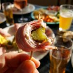 A close-up of a hand holding a single ham and pickle roll-up on a toothpick.