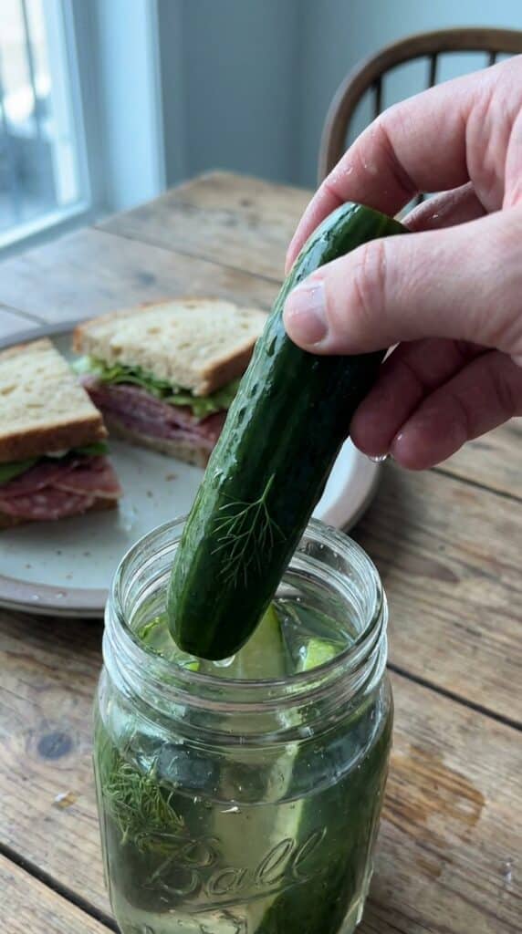 A close-up of a hand pulling a crisp, wet pickle spear from a jar, with a sandwich in the background.