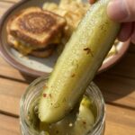 A close-up of a hand pulling a crunchy pickle spear from a jar, with a burger in the background.
