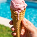 A close-up of a hand holding a waffle cone with melting strawberry ice cream in front of a sunny background.