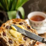A close-up of a knife spreading melting butter on a toasted hot cross bun split in half.