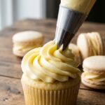 A close-up of a piping bag dispensing a smooth swirl of yellow French buttercream onto a cupcake.