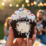 A close-up of a hand holding a bitten, melting ice cream stuffed chocolate cupcake at a backyard party.