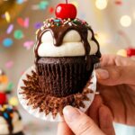 A close-up of a hand peeling the paper wrapper off an ice cream sundae cupcake, showing the moist chocolate cake and tall decorated frosting.