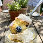 A close-up of a spoon lifting a bite of lemon, cake, and blueberry from a glass trifle cup.