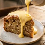 A close-up of warm vanilla custard being poured over a thick slice of Irish Apple Cake, showing the apple chunks inside.
