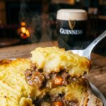 A close-up of a spoon lifting a scoop of shepherd's pie, revealing the meat and potato layers.