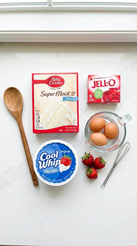 A flat lay showing a box of white cake mix, strawberry jello box, wooden spoon, and cool whip.
