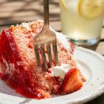 A close-up of a fork cutting into a moist slice of cake with red jello swirls and whipped cream.
