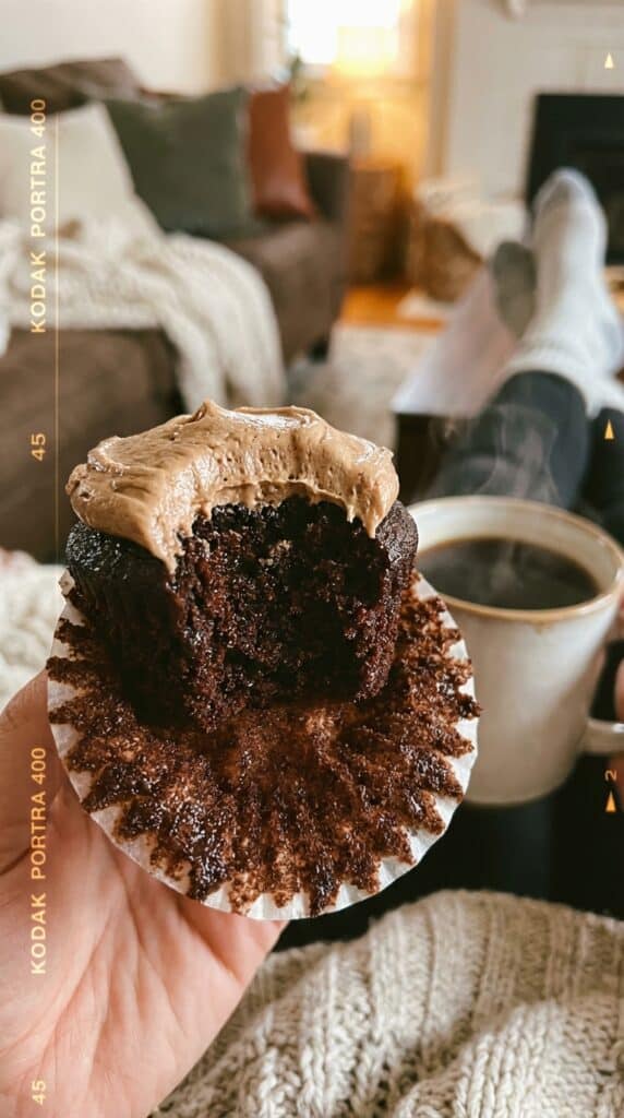 A close-up of a hand holding a chocolate Kahlúa cupcake with a bite taken out, showing the dark, moist interior crumb.