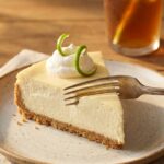A close-up of a slice of key lime cheesecake on a plate, with a fork cutting into the creamy filling.