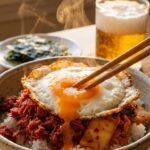 A close-up of chopsticks breaking a runny egg yolk over a bowl of beef and kimchi rice.