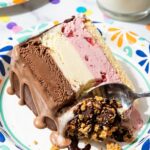 A close-up of a slice of layered ice cream cake on a plate, slightly melting, with a spoon.
