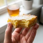 A close-up of a hand holding a lemon bar with a bite taken out, showing the texture of the filling and crust.