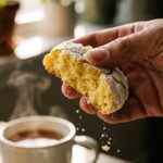A close-up of a hand breaking a soft lemon cookie in half, showing the moist interior.