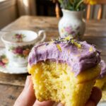 A close-up of a hand holding a bitten lemon cupcake, revealing the yellow crumb and purple frosting.