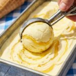 A close-up of an ice cream scoop dragging through a pan of lemon swirl ice cream, showing the ribbons of curd.