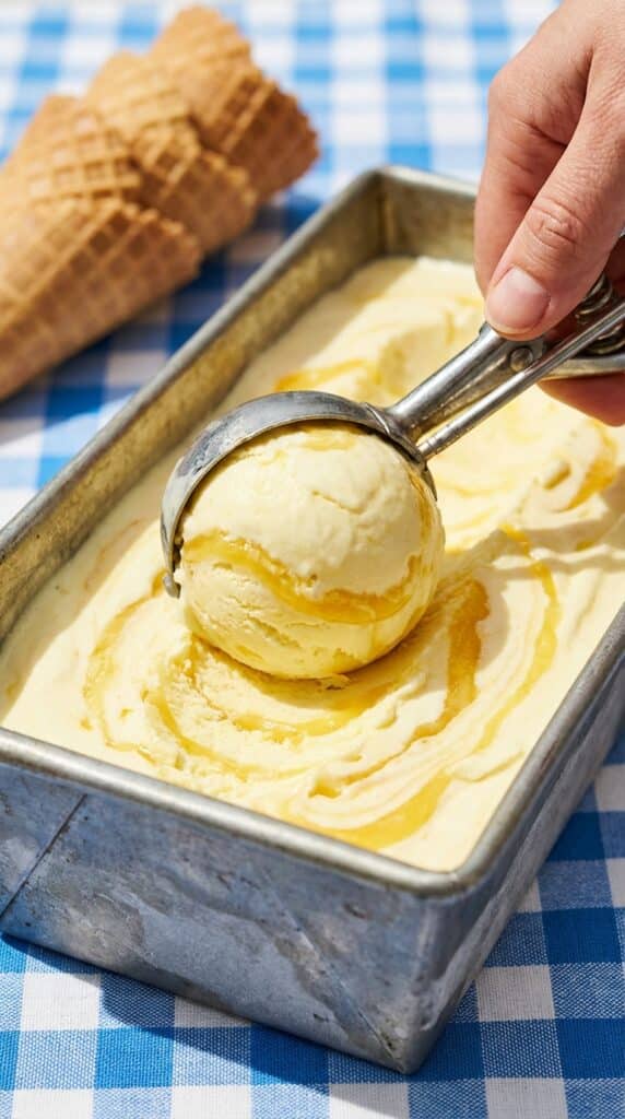 A close-up of an ice cream scoop dragging through a pan of lemon swirl ice cream, showing the ribbons of curd.