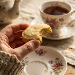A close-up of a hand holding a bitten shortbread cookie with crumbs falling, near a cup of tea.