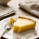 A close-up of a slice of lemon tart on a plate, showing the smooth texture and crisp crust, with a fork.