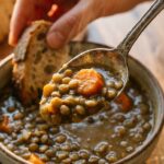 A close-up of a spoon lifting thick lentil soup from a bowl, with a piece of bread being dipped in the background.