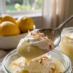 A close-up of a spoon lifting a bite of creamy lemon pudding with toasted coconut topping.