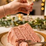A close-up of a slice of pink strawberry cake with a glass of champagne in the background.