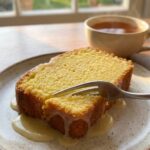 A close-up of a slice of moist lemon cake on a plate with a fork and a cup of tea.