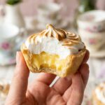 A close-up of a hand holding a mini lemon pie with a bite taken out, showing the lemon filling inside.