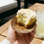A close-up of a hand holding a bitten key lime cupcake, showing the graham cracker crust layer at the bottom.