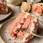 A close-up of a fork cutting into a moist slice of pink strawberry cake.