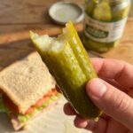 A close-up of a hand holding a bitten pickle spear, showing the crisp interior texture.