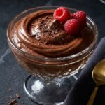 A close-up of a gold spoon lifting a thick, smooth bite of dark chocolate mousse with a raspberry.