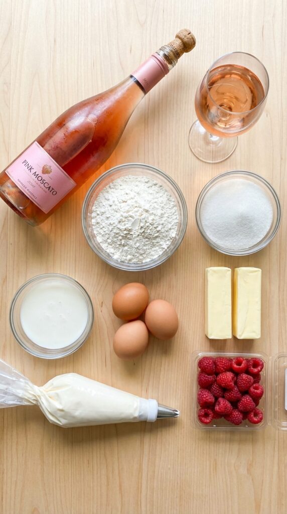 A flat lay showing a bottle of Moscato wine, flour, butter, sugar, eggs, and raspberries on a wooden table.