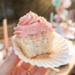 A close-up of a hand peeling the wrapper off a halved Moscato cupcake, showing the moist crumb inside.