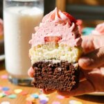 A close-up of a hand holding a cupcake cut in half, showing distinct chocolate, vanilla, and strawberry layers.