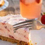 A close-up of a fork taking a bite of pink strawberry cheesecake, showing the smooth, airy texture.