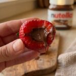 A close-up of a hand holding a bitten strawberry, showing the chocolate filling inside.