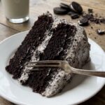 A close-up of a fork taking a bite of dark chocolate cake with speckled Oreo frosting.