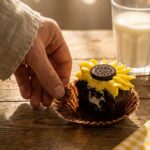 A hand peeling the wrapper off a sliced sunflower cupcake revealing a moist chocolate interior cake crumb.