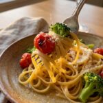 A close-up of a fork lifting a bite of creamy pasta primavera with a cherry tomato and broccoli floret, with a glass of wine in the background.