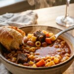 A close-up of a spoon lifting a bite of pasta e fagioli soup with bread on the side.