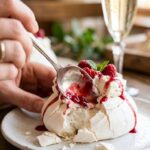 Close-up of a spoon cracking into a crisp mini pavlova, showing the soft marshmallow center, whipped cream, and red raspberry sauce.