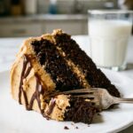 A close-up of a slice of chocolate cake with peanut butter frosting on a plate, with a fork taking a bite.