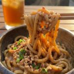 A close-up of wooden chopsticks lifting a large, saucy bite of peanut noodles from a bowl.